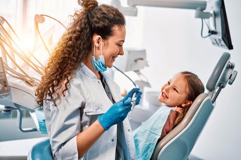 A female dentist doctor talks to a small patient, the child shows her teeth and sits in the dental chair. Sunlight.