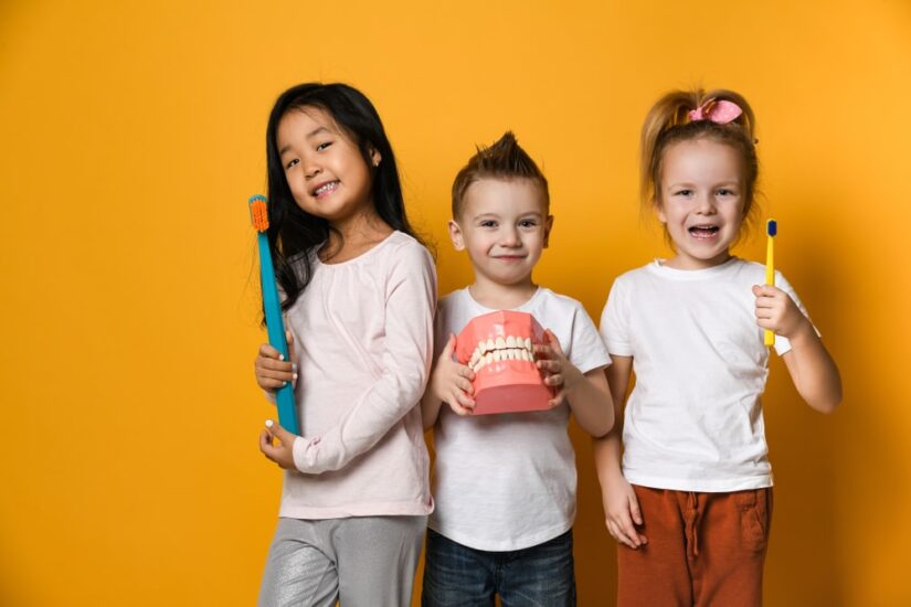 Three children with toothbrushes and a Dental implant model  stand over yellow background wall and smile rejoicing at the camera.The concept of health, oral hygiene, people and beauty.